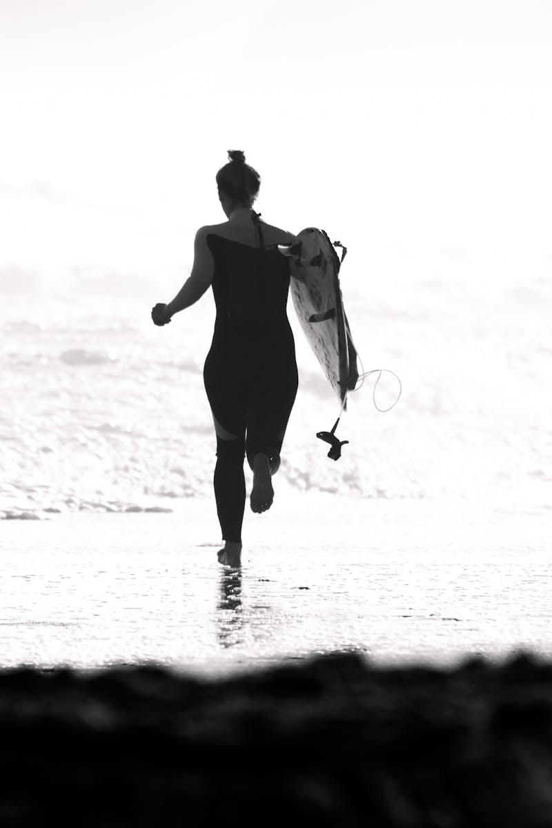 A woman carrying a surfboard runs along the beach, captured in black and white.